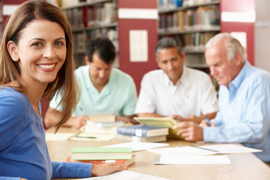 A group of adults, including one woman smiling at the camera, sit at a library table with books and papers, representing tutoring or adult education as retiree-friendly jobs.