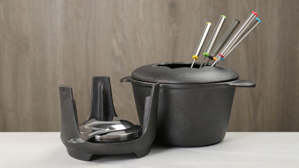 A black cast iron fondue pot with multiple stainless steel skewers sits beside a matching burner stand on a white table.