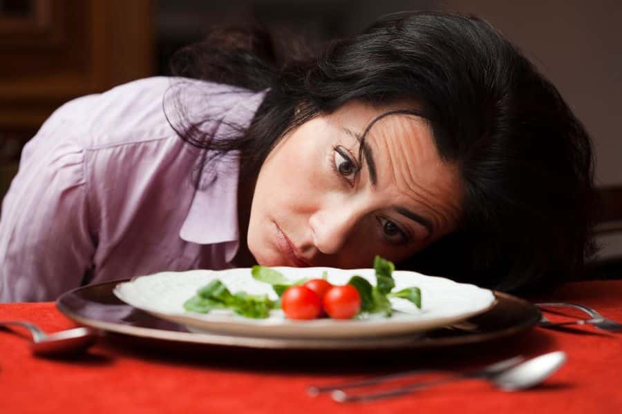 A woman rests her head on a table, looking disheartened at a plate with only two cherry tomatoes and a few greens. This image reflects the frustration many feel with restrictive diets often promoted as anti aging hacks.