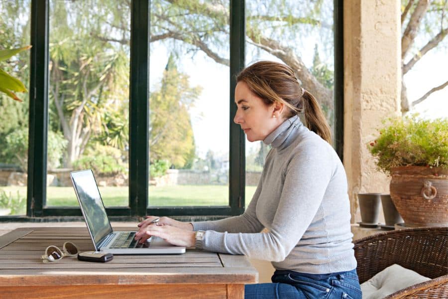 Woman in a gray turtleneck working on a laptop at a wooden table in a sunlit patio with large windows and garden views. The scene reflects a peaceful remote work setting, ideal for someone aiming to make a full-time income writing part-time.