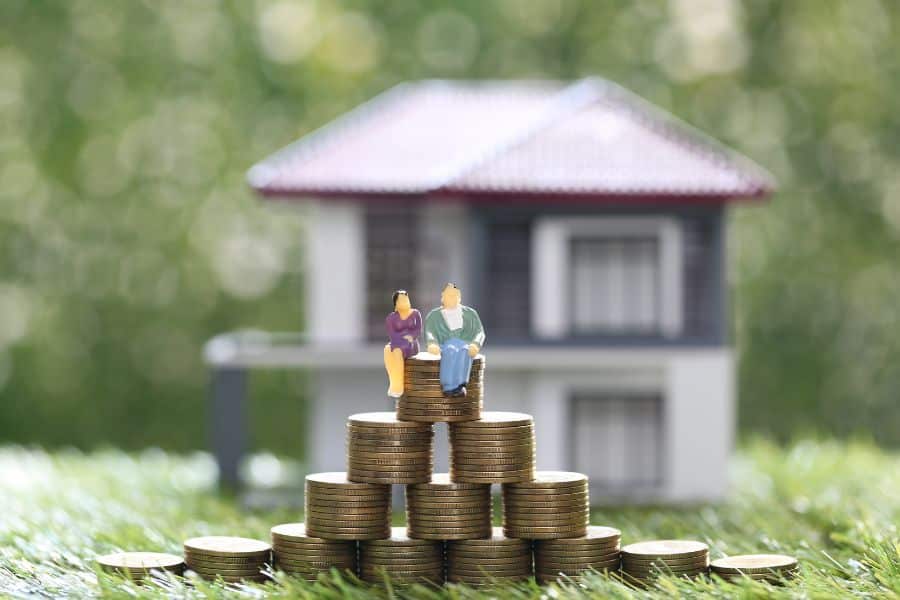 Miniature figurines of a couple sitting on stacked coins in front of a house model. Depicts saving and financial planning as a core component of long-term real estate strategies.