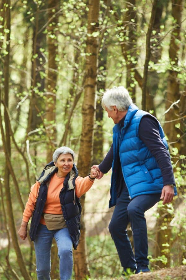 Senior couple hiking in a forest, holding hands as the man helps the woman up a slope. Hiking is a practical and bonding form of everyday exercise in nature.