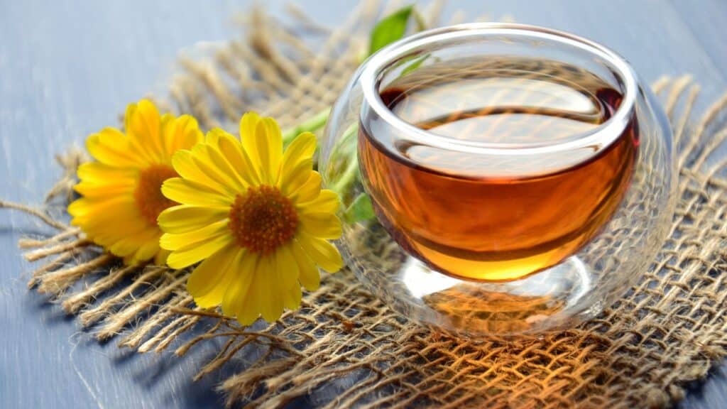 A cup of amber-colored calendula tea next to two bright yellow calendula flowers on burlap fabric. Known in vintage remedies for skin soothing and digestive support.