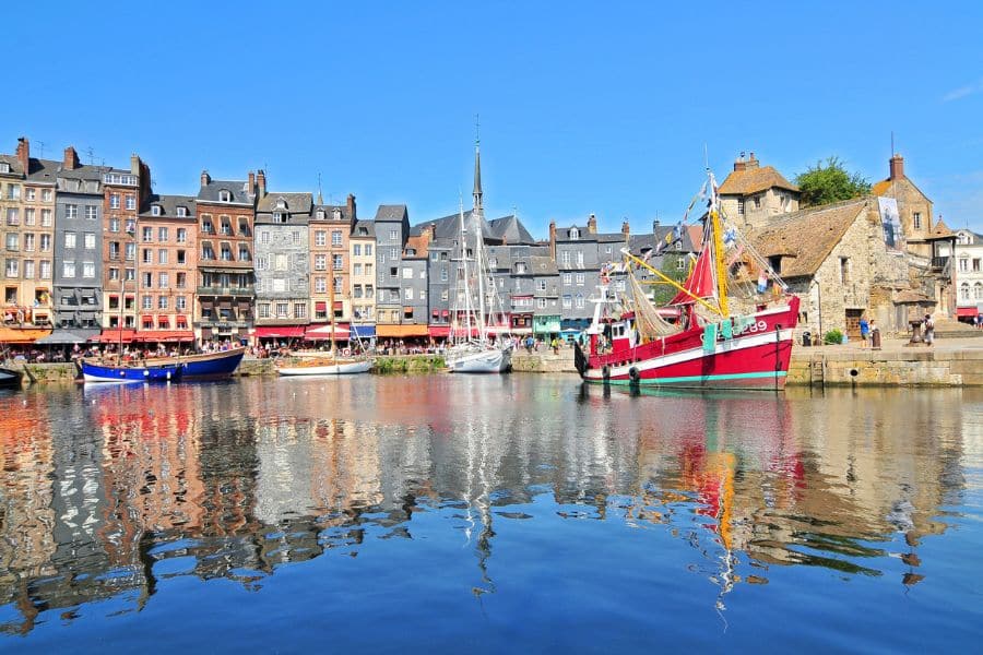 Colorful fishing boats floating in the old harbor of Honfleur, France, with tall historic buildings reflecting in the calm water. A classic northern European coastal village known for its maritime heritage.