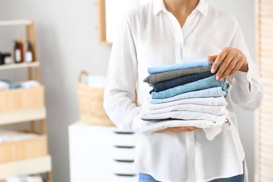 A person in a white shirt holds a neatly folded stack of clothes in a bright laundry room, representing laundry services or organizational assistance as retiree-friendly jobs.