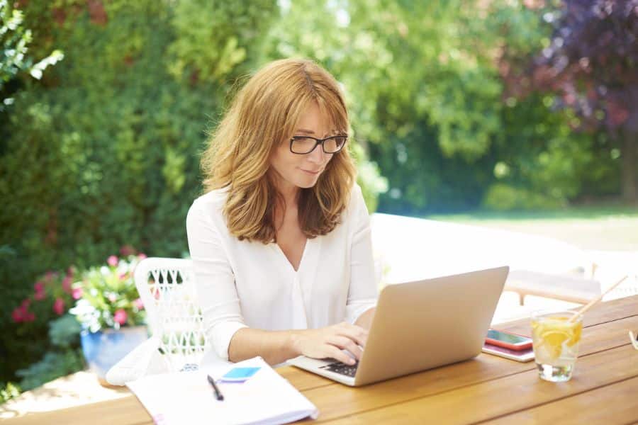 Woman with glasses typing on a laptop outdoors at a wooden table, surrounded by greenery, a drink, and open notebooks. A relaxed freelance writing setup that conveys the flexibility of making a full-time income writing part-time.