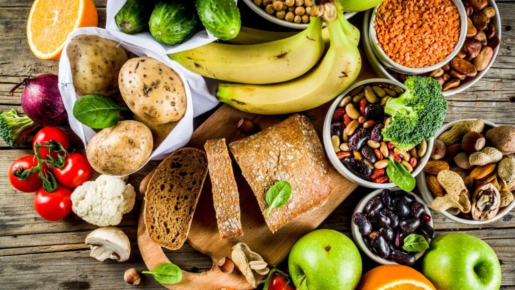 A wide assortment of plant-based foods including beans, lentils, whole grains, fruits, vegetables, and nuts on a wooden table. Great for diet tips about plant-forward nutrition and balanced meals.