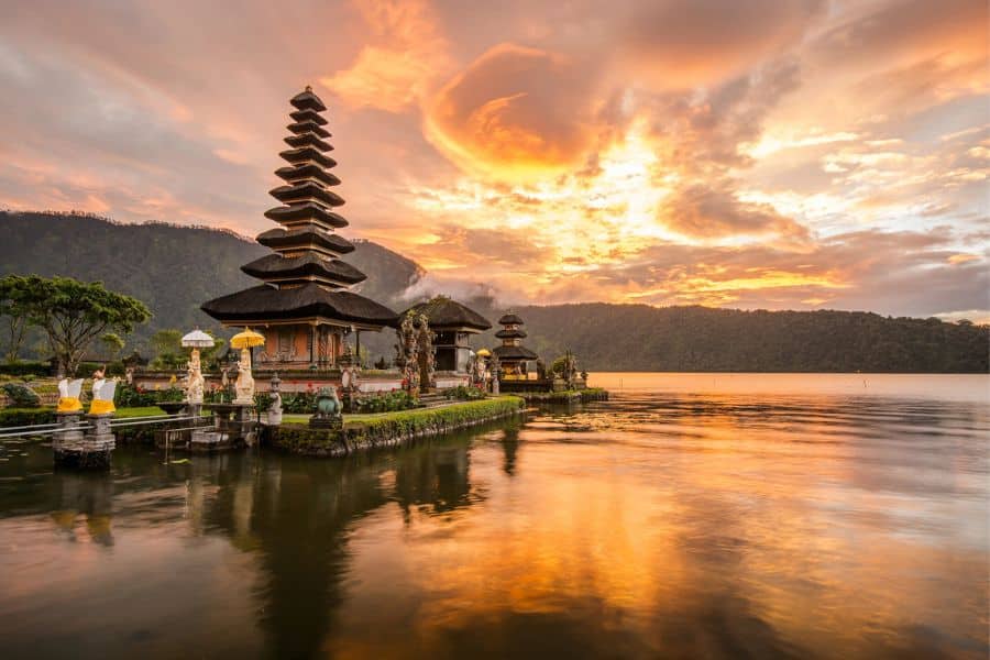 Traditional Balinese temple with multi-tiered roof perched on a small island in a lake, glowing under a dramatic orange and gold sunset sky.