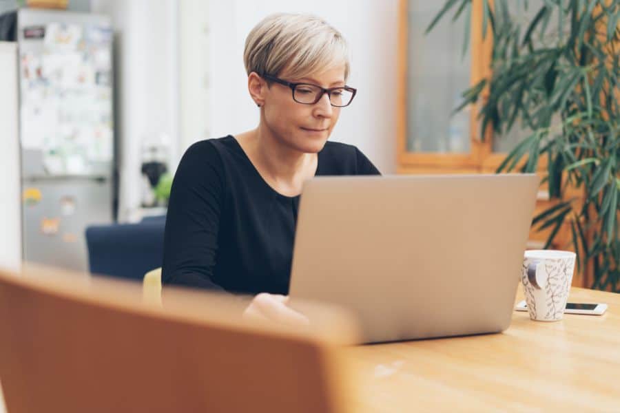 A focused woman wearing glasses working on a laptop at a wooden table, suggesting mindful productivity and strategies to beat the brain blocks.