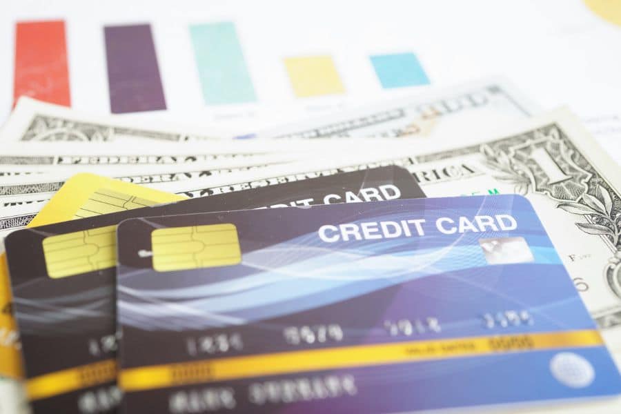 Close-up of multiple credit cards and US dollar bills on a desk with charts in the background, suggesting managing credit wisely as one of the ways to save money.