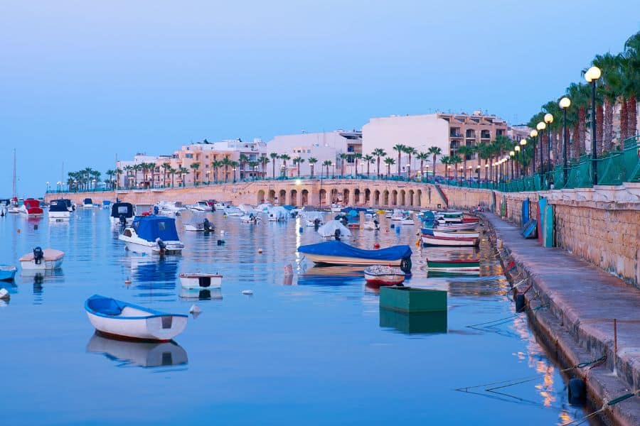 A tranquil evening view of Marsaskala, Malta, with small fishing boats floating on still water and palm-lined promenades under soft blue twilight. A peaceful coastal village atmosphere in the Mediterranean.