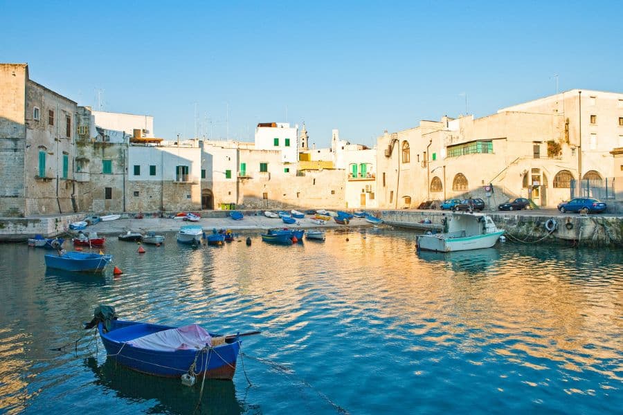 Boats docked in the small harbor of Monopoli, Italy, surrounded by whitewashed stone buildings and golden sunlight reflecting off the sea. A timeless Italian coastal village filled with rustic charm.