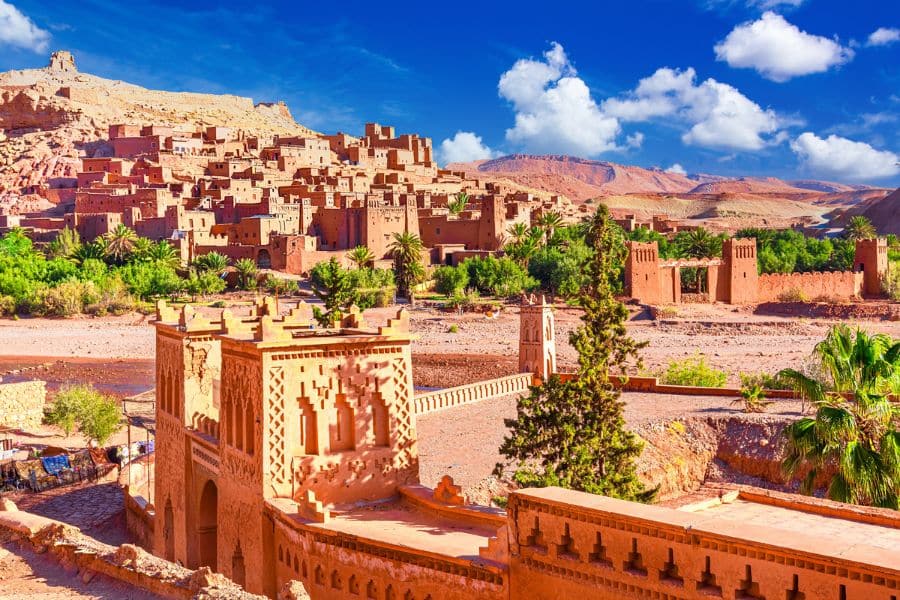 Ancient earthen clay buildings and fortified structures in the red desert landscape of Aït Benhaddou, with palm trees and blue sky in the background.