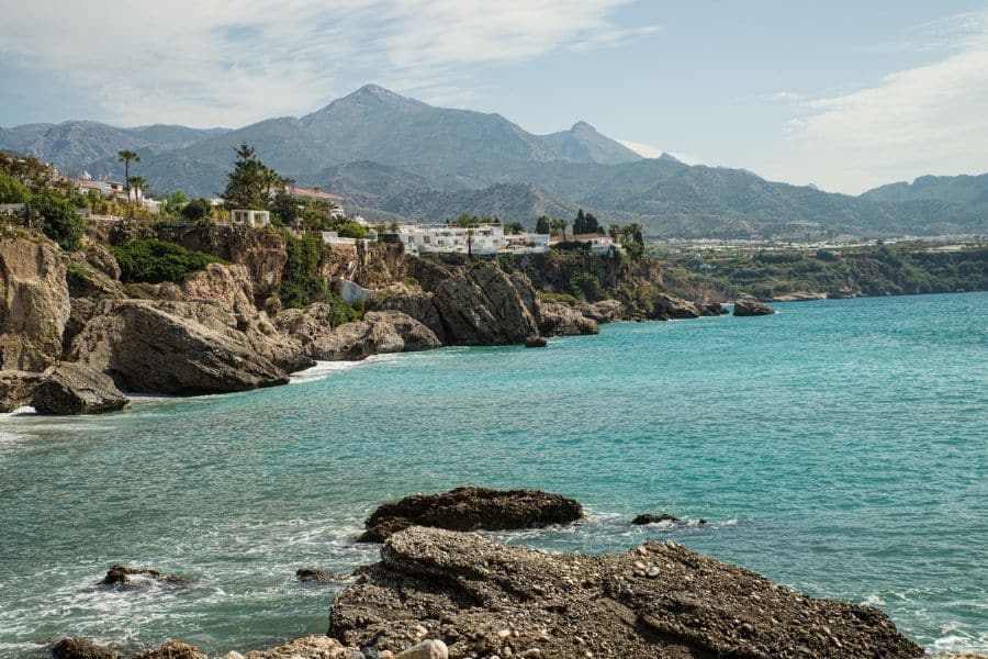 Rocky cliffs and turquoise water along the coast of Nerja, Spain, with white houses perched above and mountains in the distance. A rugged yet scenic view from one of Spain’s coastal villages.