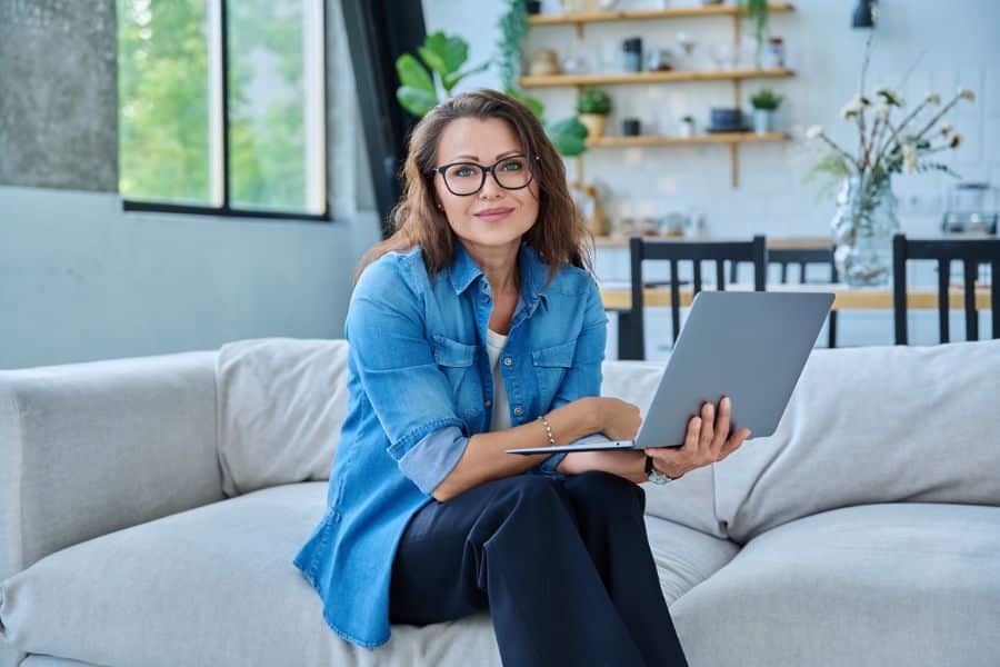 A woman sits comfortably on a couch using a laptop, illustrating freelance or remote work as a retiree-friendly job from home.