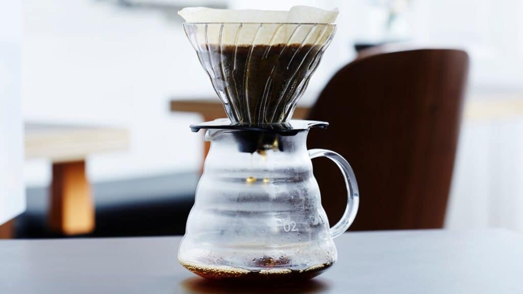 A clear glass pour-over coffee maker with a paper filter and fresh grounds, slowly brewing coffee into the lower carafe on a dark table.