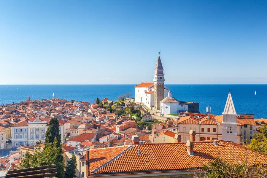 Aerial view of Piran, Slovenia, with terracotta rooftops, a prominent church tower, and the Adriatic Sea stretching into the horizon. A classic coastal European village with medieval roots.