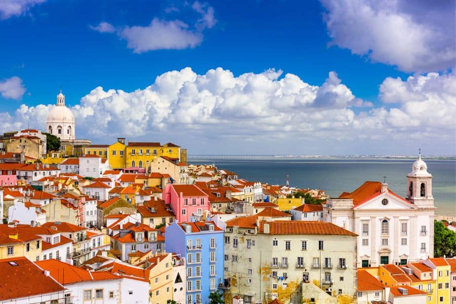 Colorful buildings with red-tiled roofs in Lisbon's Alfama district, with a domed church and ocean visible under a bright blue sky.