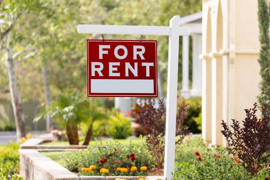 Red and white “For Rent” sign in front of a landscaped residential home. Highlights rental property strategies for generating passive income in real estate.