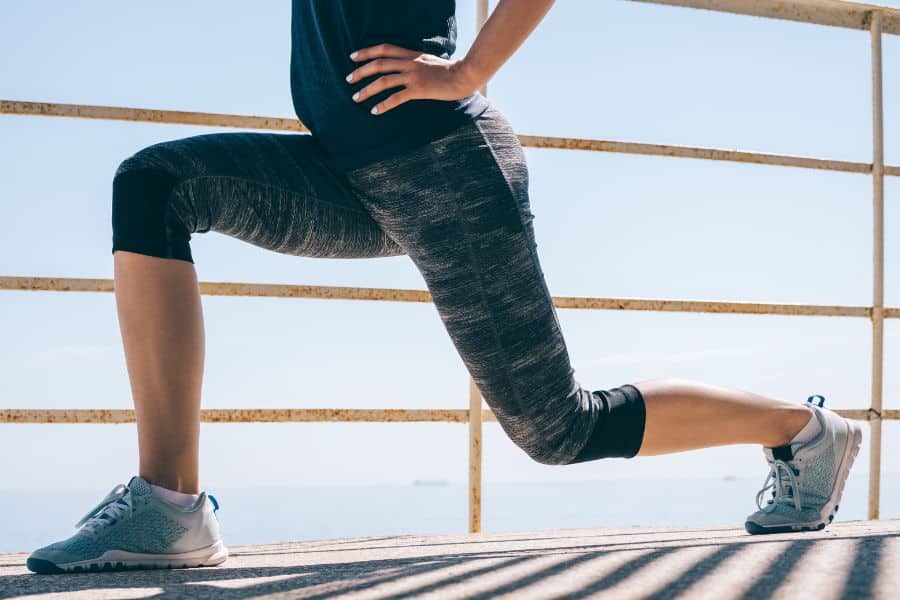 Close-up of a person lunging outdoors on a sunlit deck with hands on hips and ocean in the background. Demonstrates a simple standing exercise suitable for building leg strength.