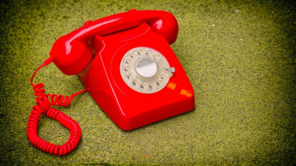 A bright red rotary phone with its curly cord placed on green textured outdoor flooring, giving off retro emergency phone vibes.