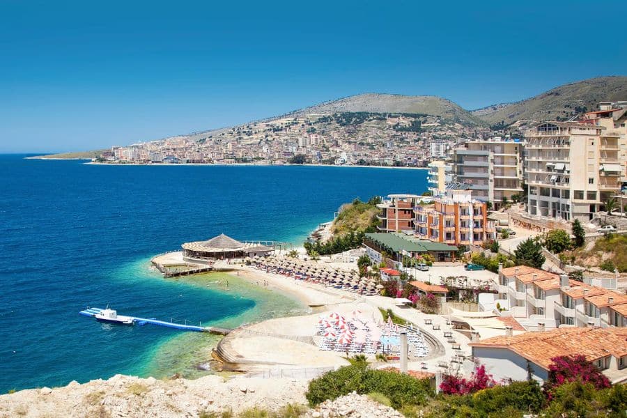 A panoramic view of Sarandë, Albania, showing a curved beach with umbrellas, turquoise water, and hillside buildings overlooking the Ionian Sea. A rising star among European villages on the coast with a vibrant, resort-like charm.