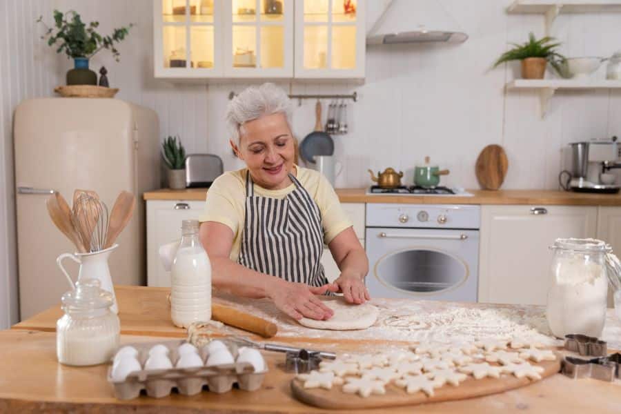 An older woman in a striped apron kneads dough in a cozy kitchen surrounded by baking ingredients, illustrating home baking or cottage food sales as retiree-friendly jobs.