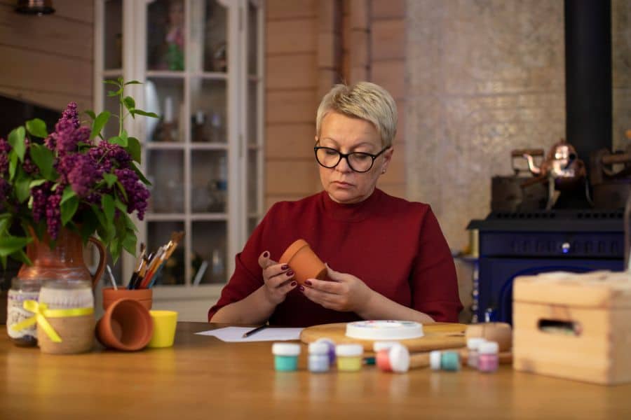 A woman with short blonde hair examines a small terracotta pot with paint jars on the table, showcasing crafting or handmade goods sales as retiree-friendly job ideas.