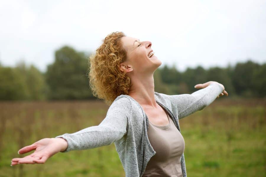 A joyful woman stands in a grassy field with arms wide open, eyes closed, and face tilted toward the sky. Embracing nature, fresh air, and stress relief can be some of the simplest and most powerful anti aging hacks.