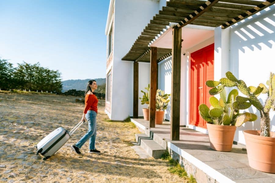 Woman with a suitcase walking toward a modern vacation rental with red doors and large potted cacti. Represents short-term rental strategies in real estate investment.