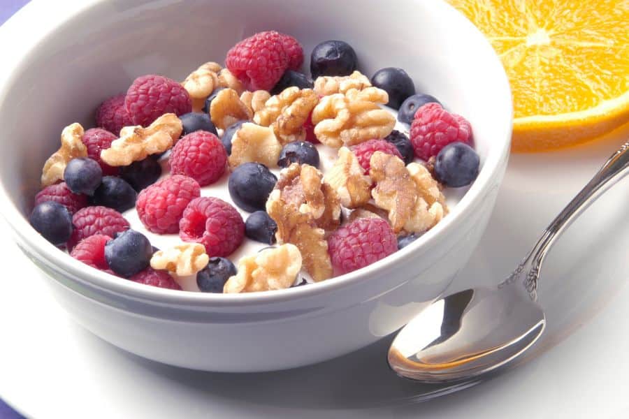 A bowl of yogurt topped with raspberries, blueberries, and walnuts, with a slice of orange on the side, showcasing brain-boosting foods that help beat the brain blocks.