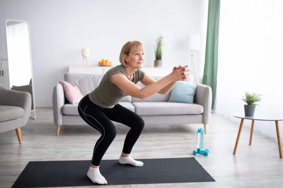 Mature woman doing a squat on a yoga mat in a cozy living room with her arms extended forward. A gentle standing workout great for beginners or at-home routines.