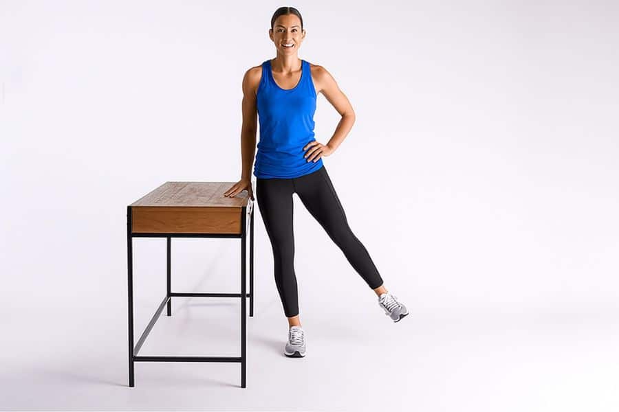 Smiling woman in Black and blue athletic wear stands beside a table while lifting one leg to the side. This balance-based move is a classic example of an easy standing exercise.