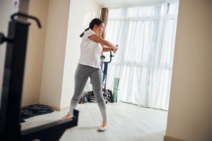 Young woman in grey leggings and white shirt twists her torso while standing in a home gym. A low-impact standing core exercise that fits well in easy standing workouts.