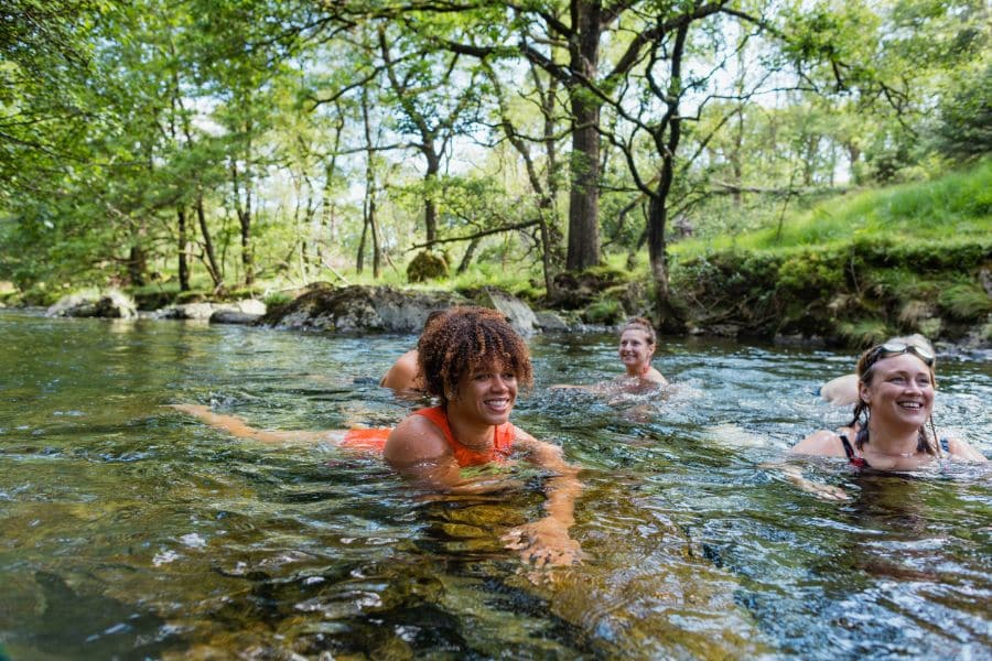 Three women smile while swimming in a clear, forested river surrounded by trees and rocks. Cold water immersion and outdoor movement are emerging as natural anti aging hacks.