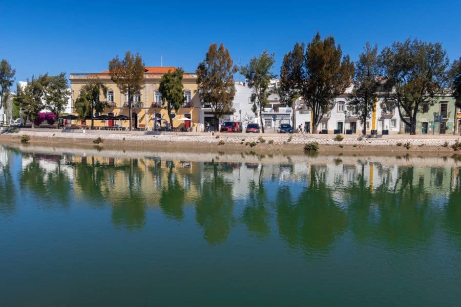 A calm riverfront in Tavira, Portugal, with pastel houses, outdoor cafés, and leafy trees mirrored in the still water. A peaceful coastal village atmosphere that reflects the slow pace of southern European life.