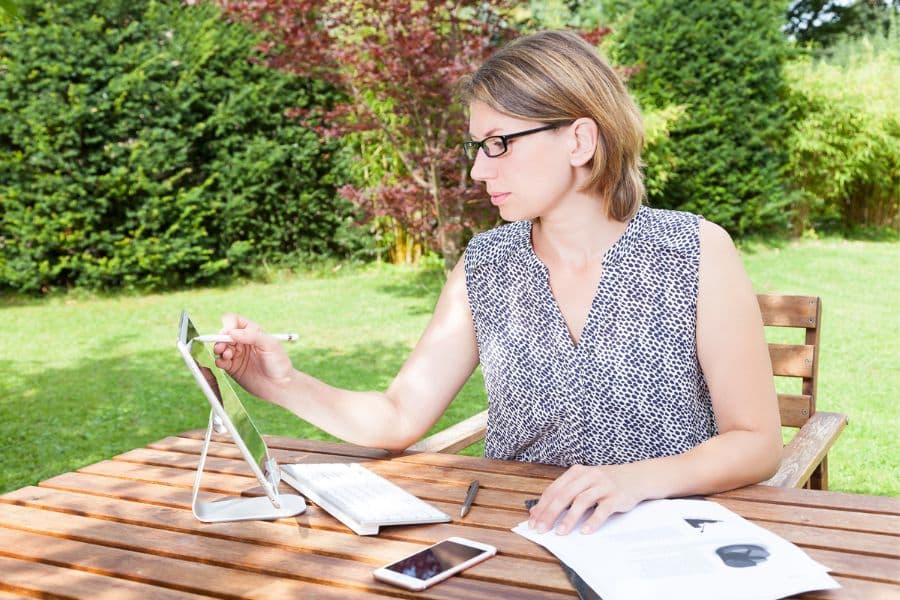 Woman using a stylus with a tablet at a patio table, surrounded by papers and a smartphone, with a lush green garden in the background. A calm and focused workspace that illustrates the potential to make a full-time income writing part-time.