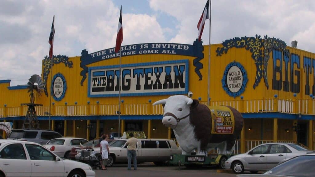 Bright yellow and blue exterior of The Big Texan Steak Ranch with Texas flags, bold signage, and a giant steer statue near the entrance. A famous Route 66 restaurant known for its 72-ounce steak challenge.