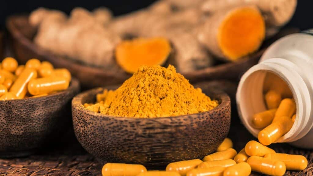 A rustic wooden bowl filled with vibrant turmeric powder, surrounded by turmeric root slices and yellow capsules spilling from a bottle. Turmeric is widely used in vintage remedies for inflammation and immune support.