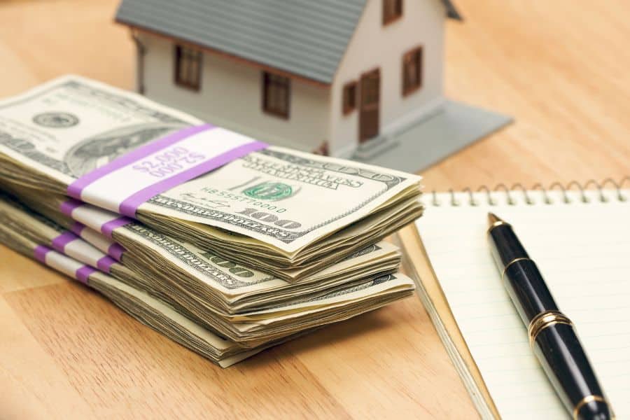 Stacks of hundred-dollar bills next to a house model, pen, and notepad on a wooden table. Conveys capital investment and budgeting as foundational real estate strategies.