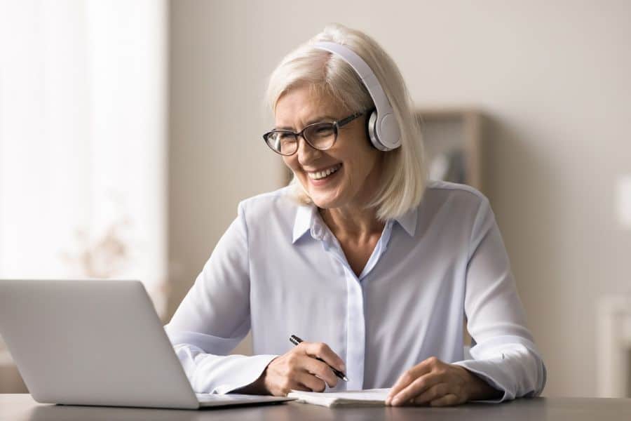 A smiling older woman wearing headphones takes notes while working on a laptop, representing online tutoring or remote consulting as ideal retiree-friendly jobs.