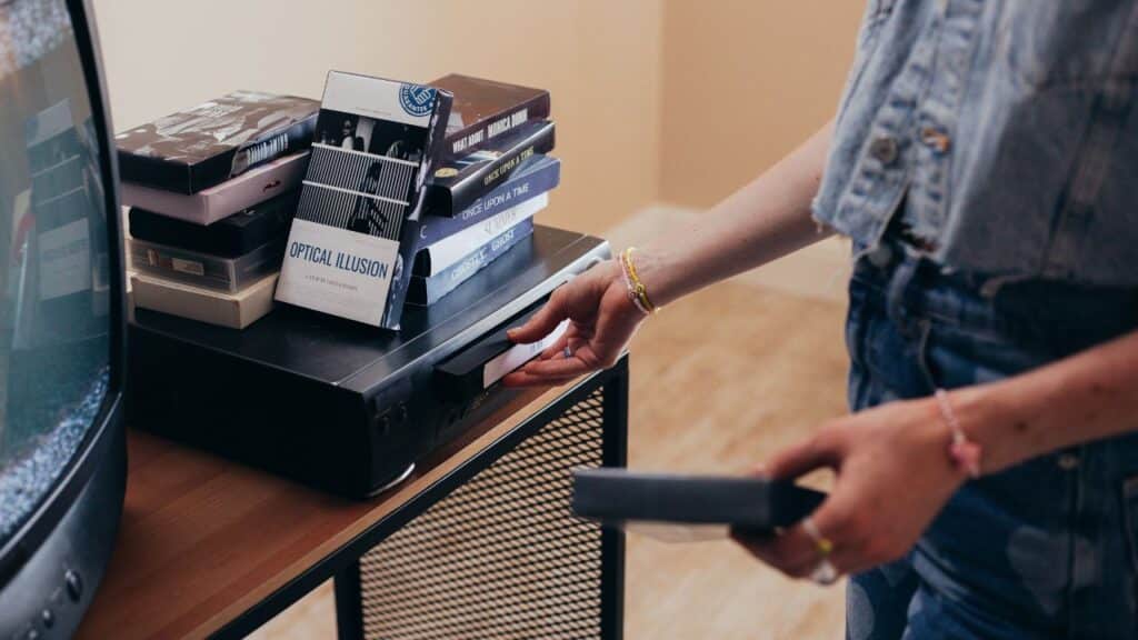 A person inserting a VHS tape into a VCR with a stack of other VHS tapes labeled with movie titles next to a tube TV.