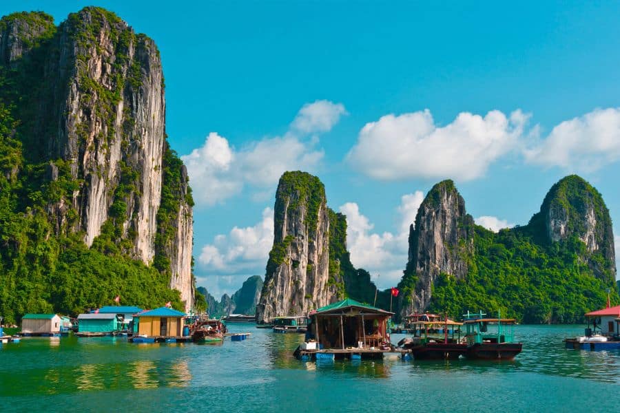 Floating fishing village and boats nestled among towering limestone karsts covered in greenery, set against a bright blue sky and calm bay waters.