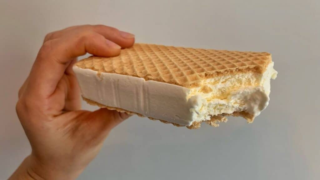 Close-up of a hand holding a vanilla ice cream sandwich with crisp waffle-style wafers, one bite taken from the side.