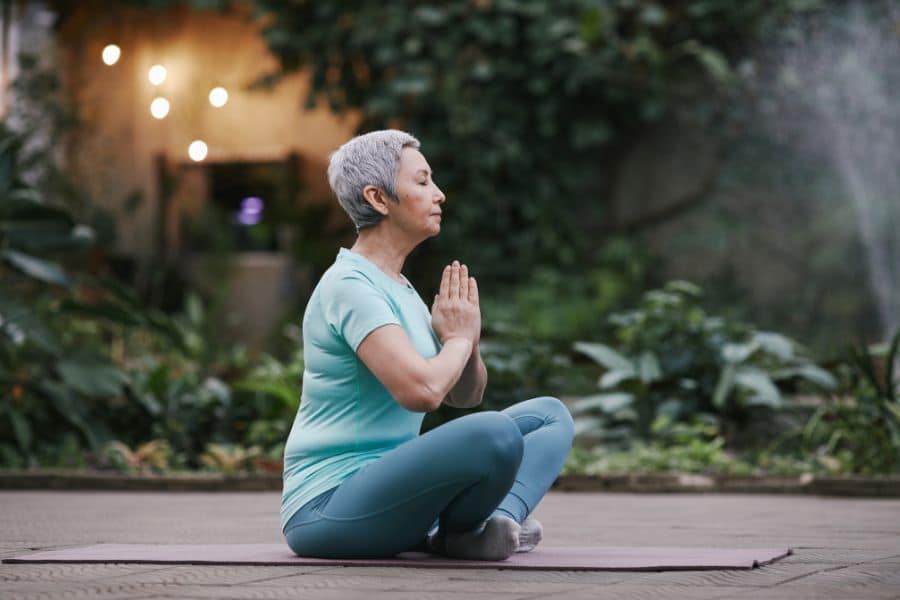 Older woman sitting cross-legged on a yoga mat outdoors, hands in prayer position, eyes closed. Yoga as a calming and restorative everyday exercise for balance and mental clarity.