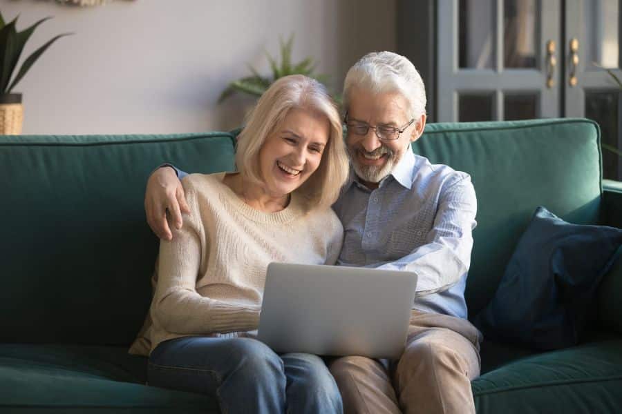 An older couple sitting closely on a green sofa, smiling while looking at a laptop together. The relaxed and joyful moment captures the lifestyle of happy American retirees engaging with technology at home.
