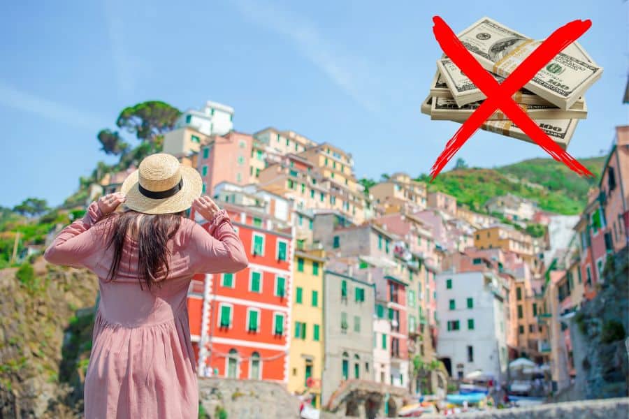 A woman in a pink dress and straw hat admires the colorful cliffside buildings of Cinque Terre, Italy, with a red "X" over a bundle of cash, symbolizing budget travel hacks.