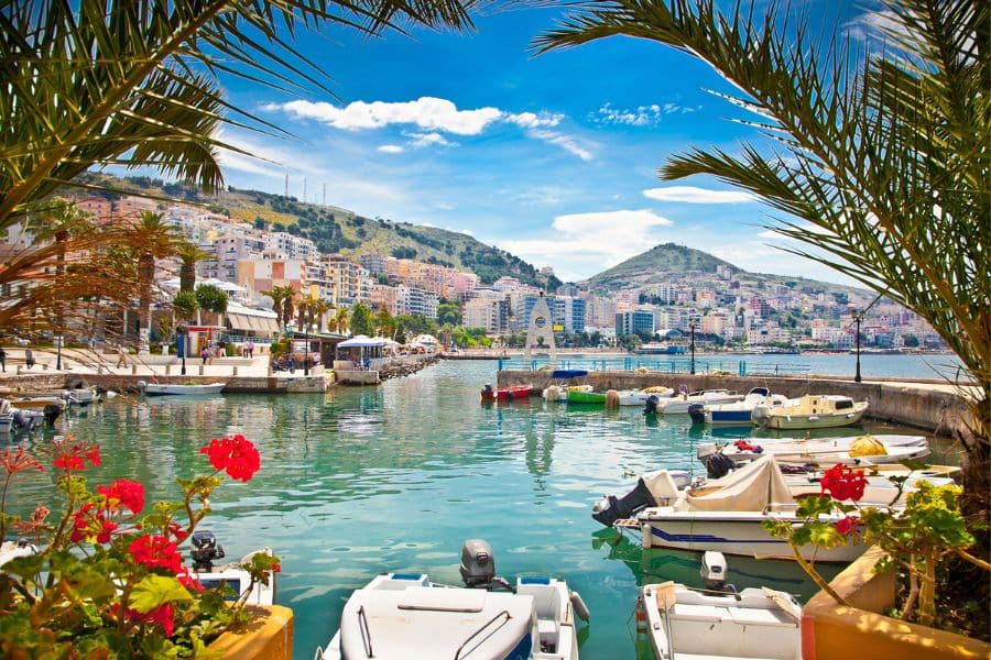 Scenic view of a coastal marina in Albania framed by palm fronds and red flowers, with colorful hillside buildings and small boats docked in turquoise waters. Ideal visual for promoting American travel to Albania’s vibrant seaside towns.