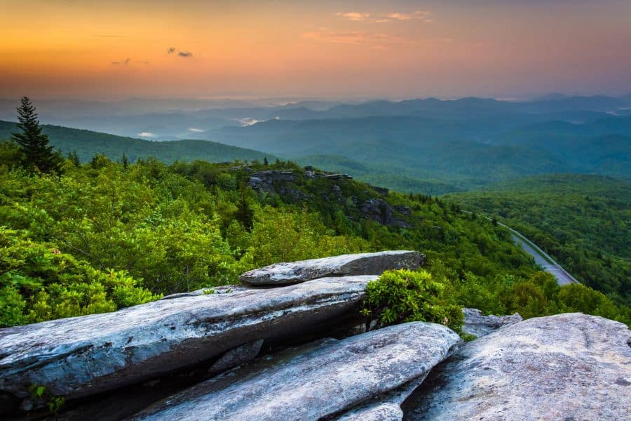 A scenic overlook reveals rolling green mountains at sunrise, with rocky ledges in the foreground and a winding road below. While not wintery, this stop could be part of a varied winter RV trip that includes milder southern destinations.