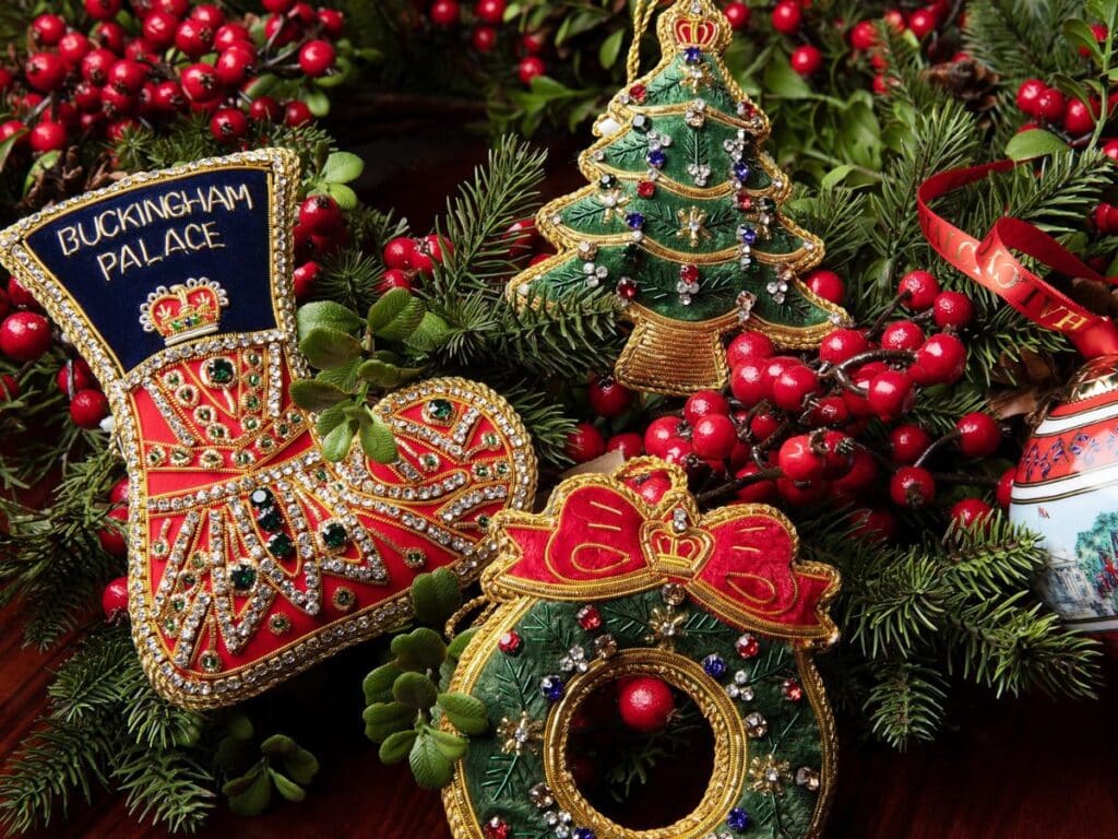 Close-up of intricate embroidered Christmas ornaments including a stocking labeled “Buckingham Palace,” a jeweled wreath with a red bow, and a sparkling Christmas tree, all surrounded by pine branches and red berries. These luxury decorations reflect the regal flair found in European Christmas markets.